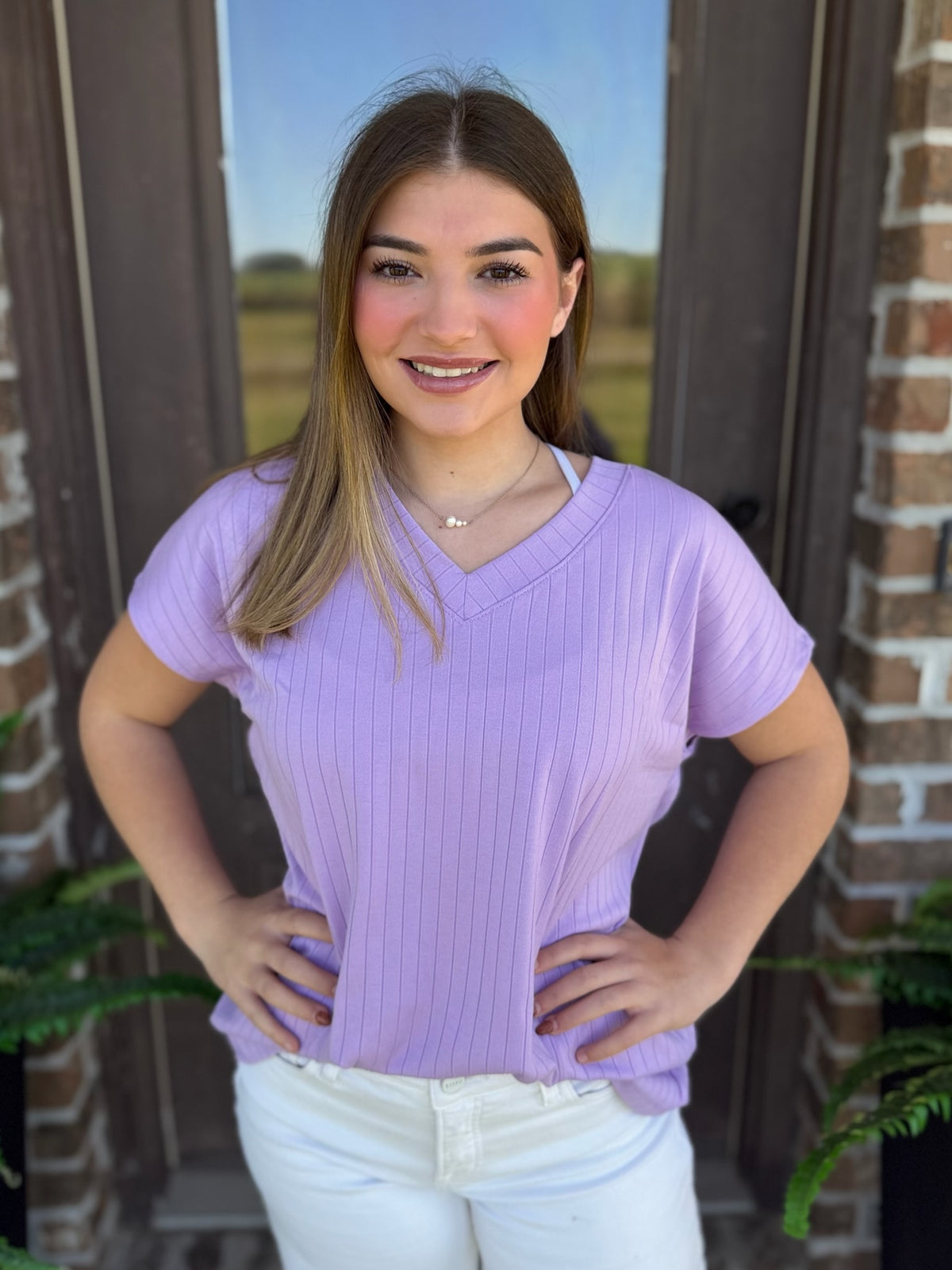 A woman wearing a ribbed lavender V-neck top with short sleeves stands in front of a brick wall.