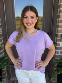 A woman wearing a ribbed lavender V-neck top with short sleeves stands in front of a brick wall.