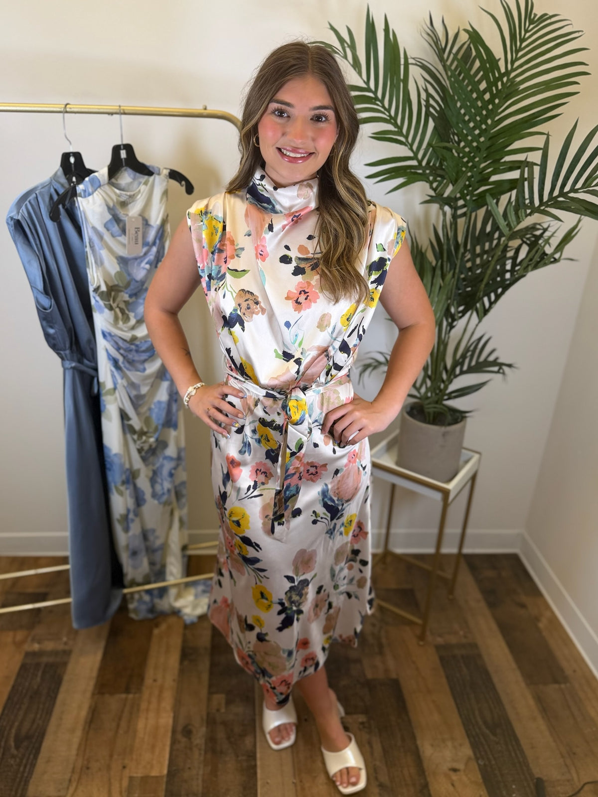 Woman in a multicolor floral midi dress standing in a room with clothing racks and plants.