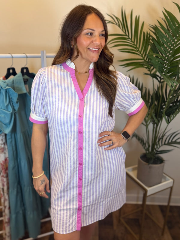 Woman wearing lavender and white striped dress with short sleeves and pink and white trim details in a clothing store setting.