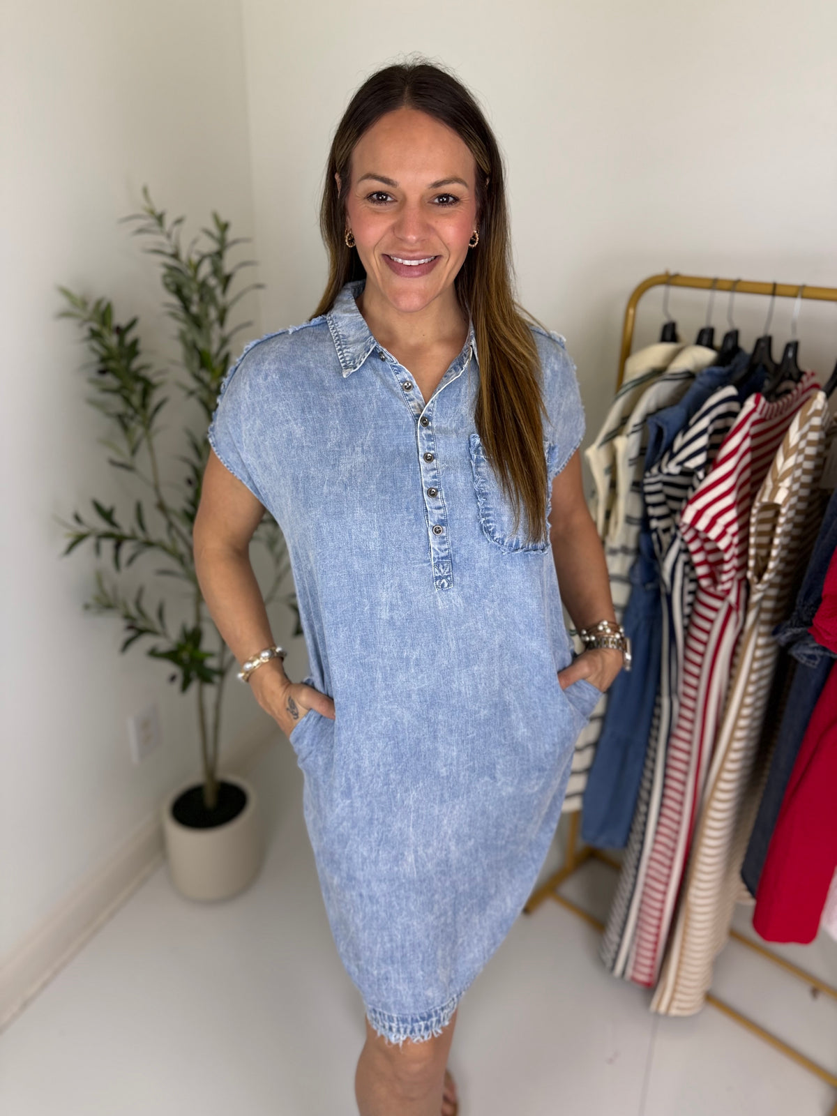 Woman wearing a light wash denim mini dress with short sleeves and a collared neckline standing next to a clothing rack with various garments.