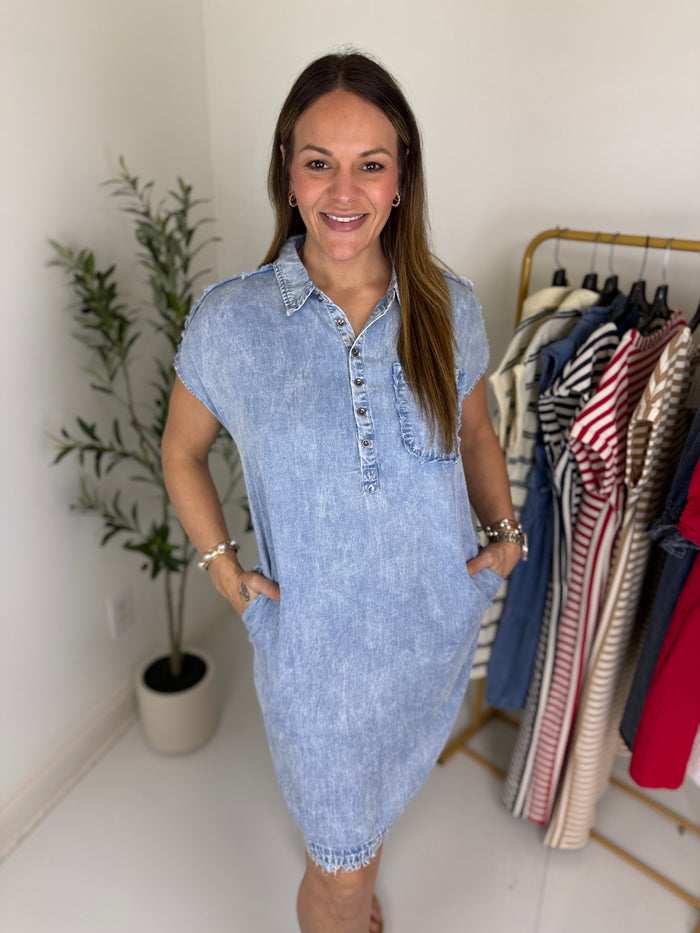 Woman wearing a light wash denim mini dress with short sleeves and a collared neckline standing next to a clothing rack with various garments.