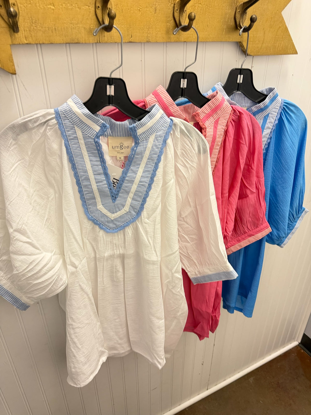A white short-sleeve blouse with a light blue striped V-neck trim hanging on a black hanger, with pink and blue blouses behind it on a white paneled wall.