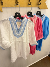 A white short-sleeve blouse with a light blue striped V-neck trim hanging on a black hanger, with pink and blue blouses behind it on a white paneled wall.