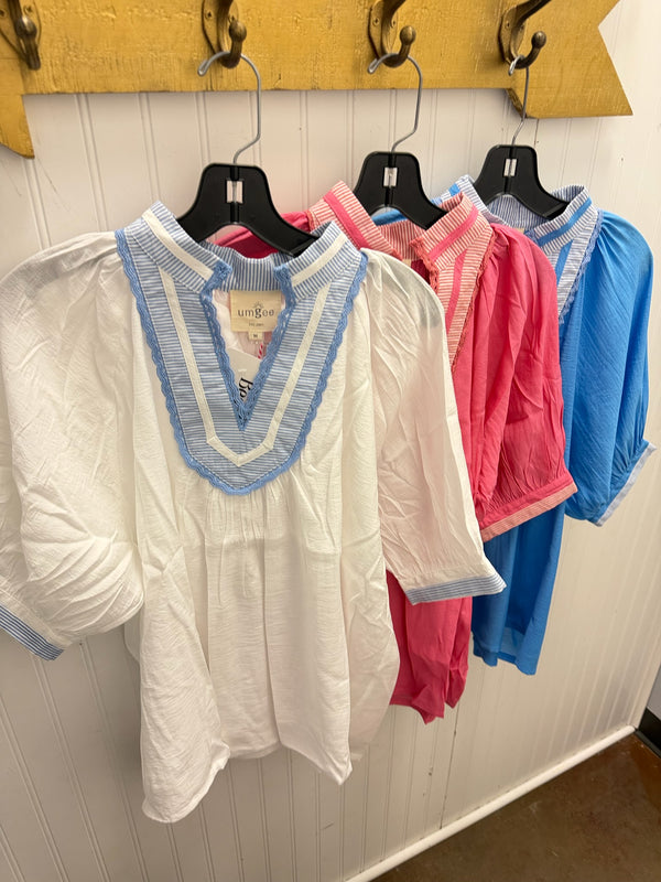 A white short-sleeve blouse with a light blue striped V-neck trim hanging on a black hanger, with pink and blue blouses behind it on a white paneled wall.
