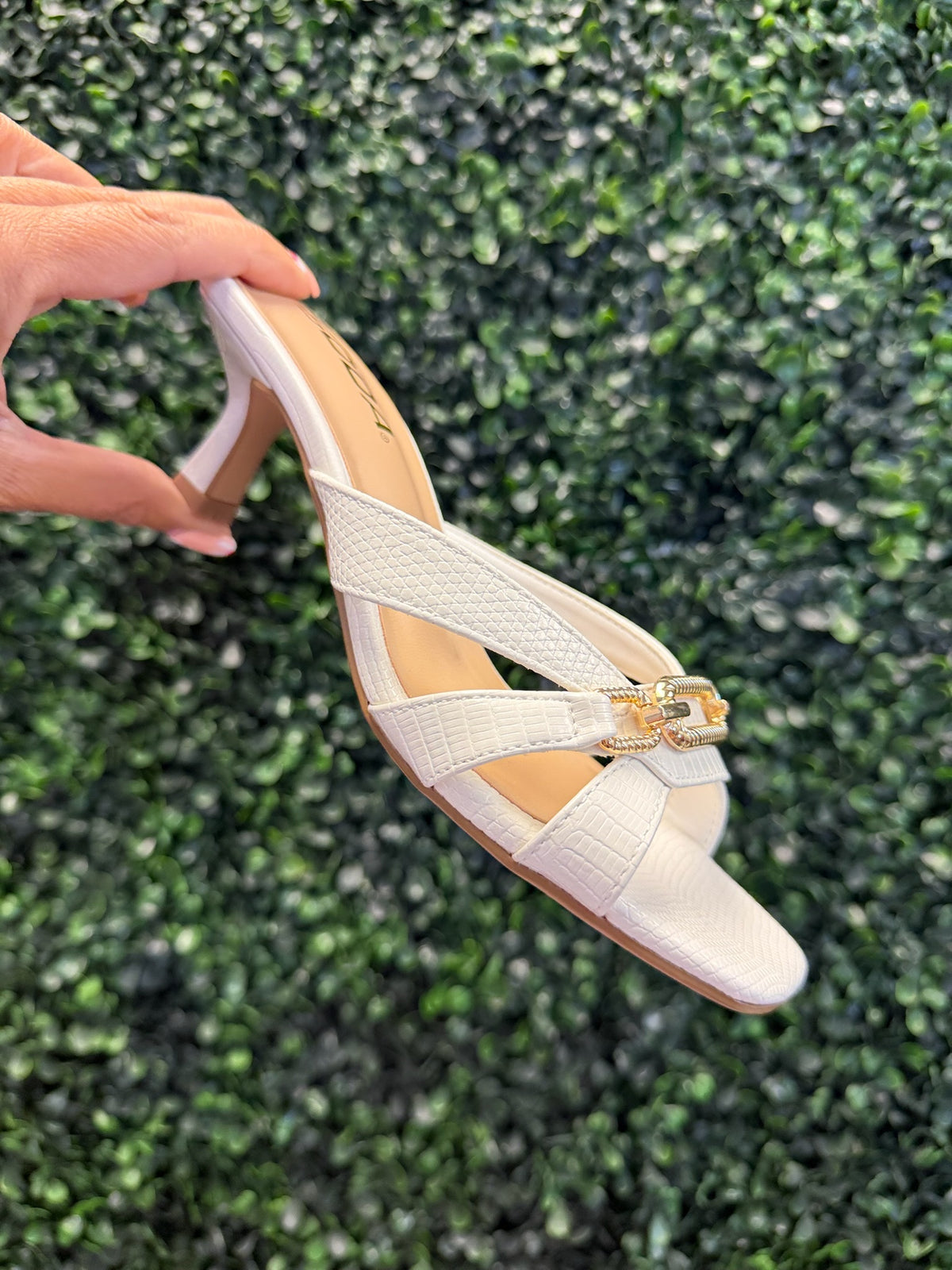 White high heeled sandal with a gold strap detail held against a green textured background