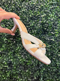 White high heeled sandal with a gold strap detail held against a green textured background