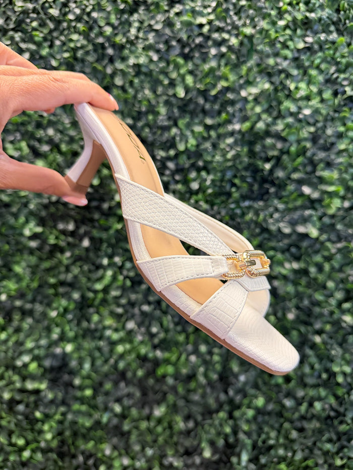 White high heeled sandal with a gold strap detail held against a green textured background