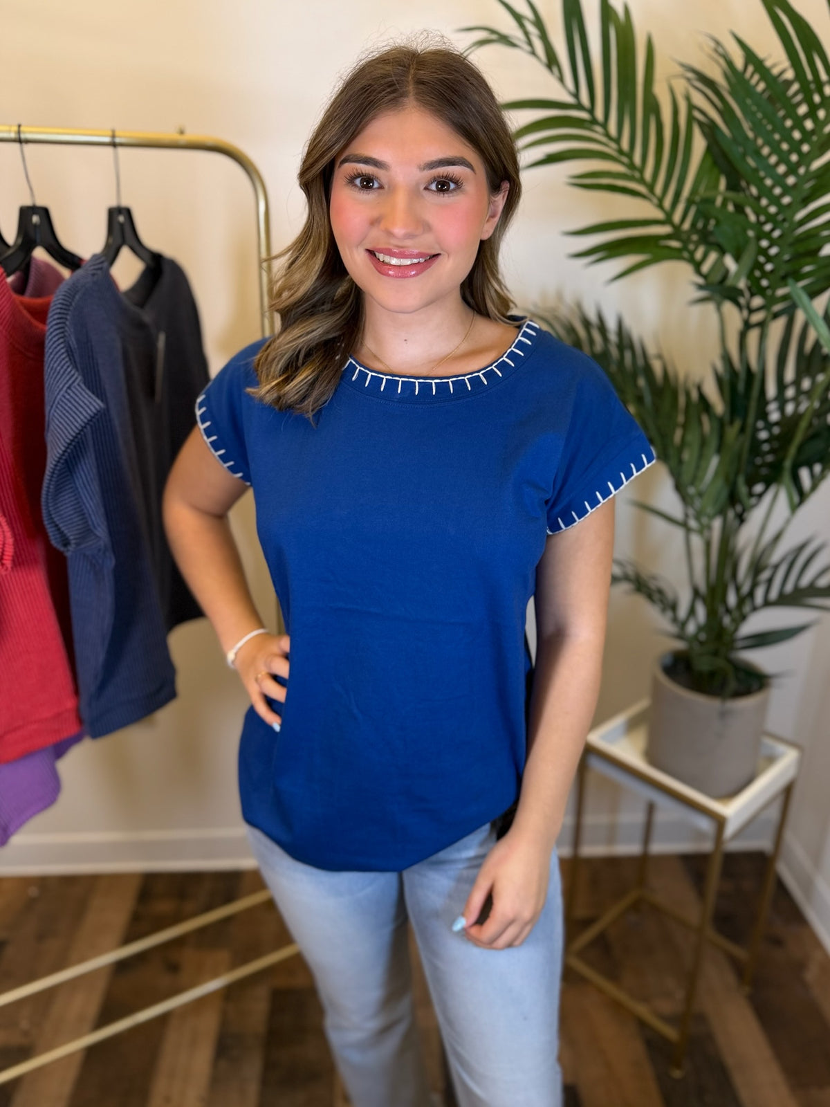 A woman modeling a royal blue short-sleeve top with white stitched trim around the neckline and sleeve cuffs, paired with light blue jeans in a boutique setting with a clothing rack and potted plant.
