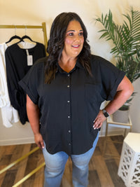 A woman modeling a black short-sleeve button-up shirt with light blue jeans in a boutique setting with clothing racks and a plant in the background.