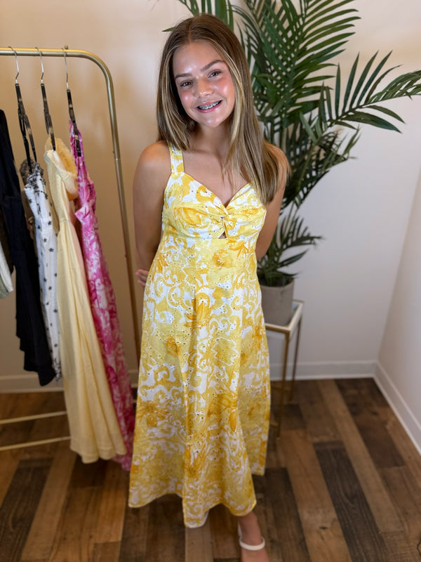 Woman wearing a sleeveless yellow floral eyelet dress standing in a room with a rack of clothes and a plant.