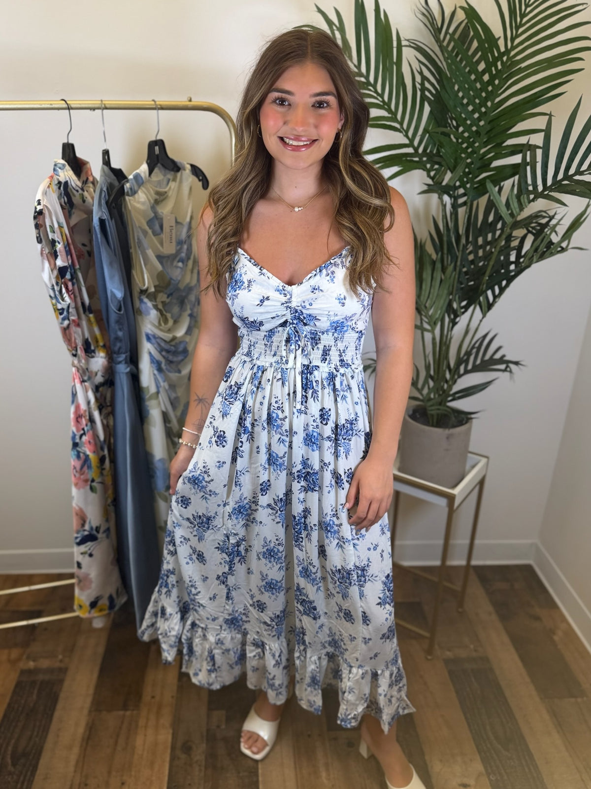 Woman wearing a white and blue floral maxi dress standing in a room with a rack of dresses and a plant.