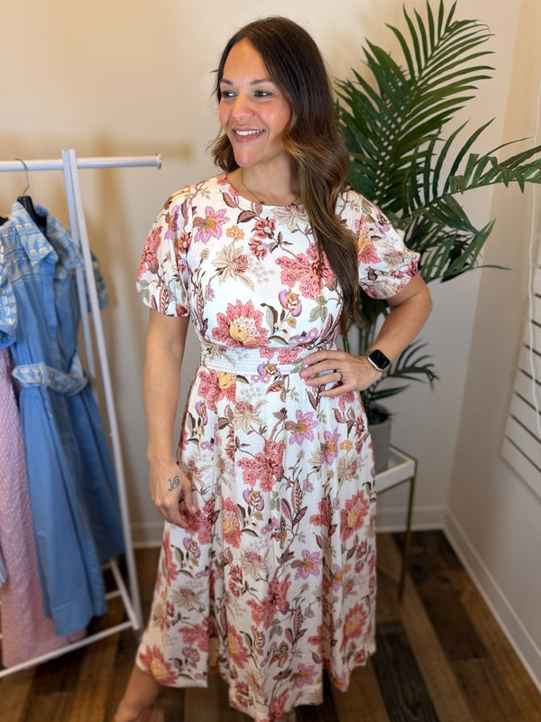 Woman wearing a floral dress midi dress with round neck and puff sleeves standing in a room with a plant and clothing rack.