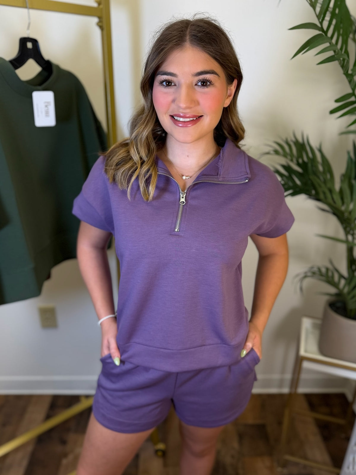 A woman modeling a matching purple short-sleeve quarter-zip polo shirt and shorts set in an indoor boutique setting with a clothing rack and plant visible in the background.