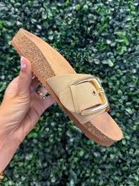Beige platform slide sandal with a gold buckle held against a green leafy background