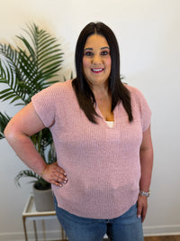 Woman wearing a pink short-sleeve sweater standing indoors with a plant in the background
