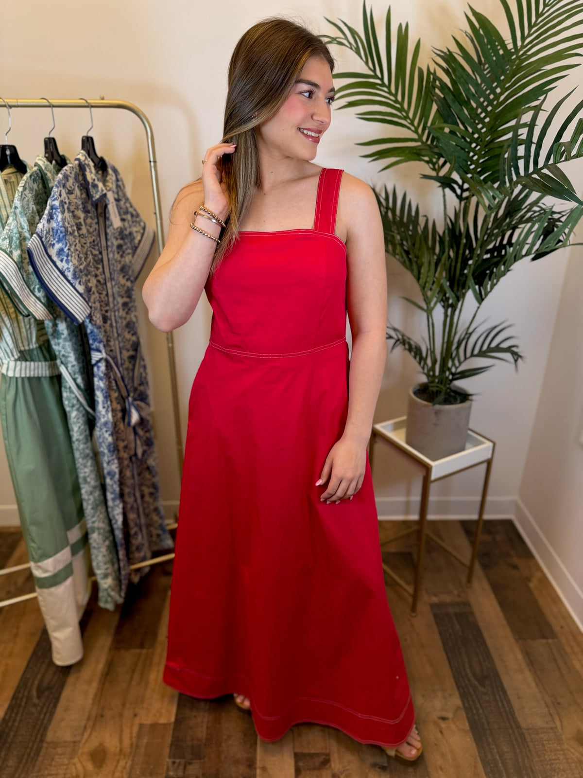 Woman in a red sleeveless maxi dress standing in a room with clothing racks and plants.