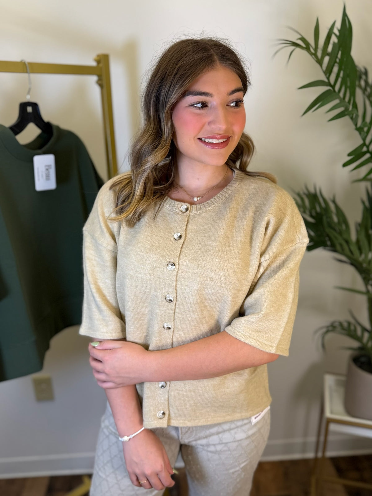 A woman modeling a short-sleeve knit top in a warm beige color with a button-down front, standing in an indoor boutique setting with a clothing rack and plant visible in the background.