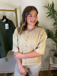 A woman modeling a short-sleeve knit top in a warm beige color with a button-down front, standing in an indoor boutique setting with a clothing rack and plant visible in the background.