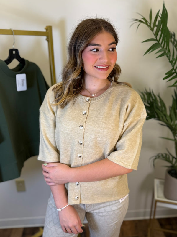 A woman modeling a short-sleeve knit top in a warm beige color with a button-down front, standing in an indoor boutique setting with a clothing rack and plant visible in the background.