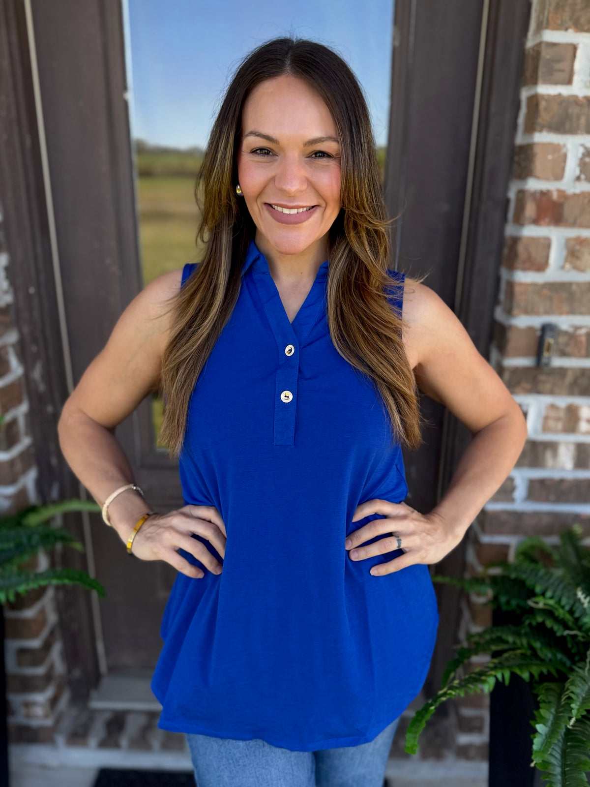A woman wearing a sleeveless royal blue polo-style top with a three-button placket, standing in front of a brick doorway.