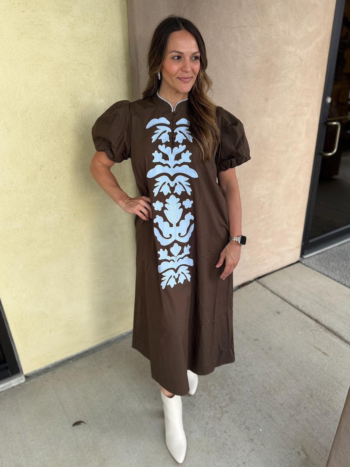 A woman wearing a brown midi dress with puff sleeves and a vertical light blue damask pattern on the front, paired with white ankle boots, standing outdoors against a beige wall.