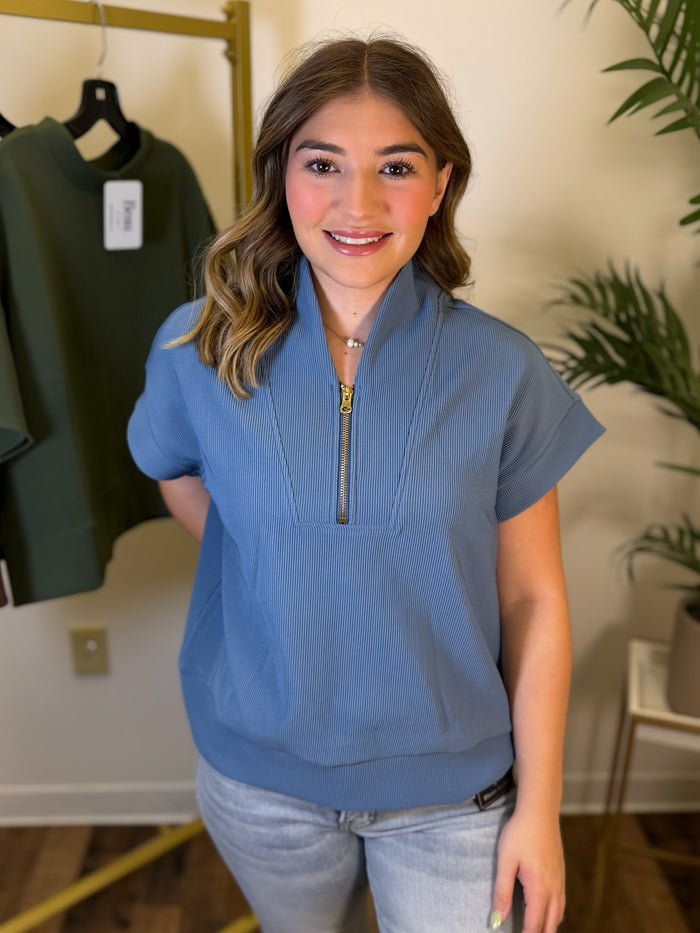 A woman wearing a dusty blue short-sleeve mock-neck top with a half-zip collar and ribbed cuffs, paired with light-wash jeans, standing in an indoor boutique setting.