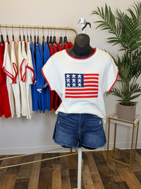 Mannequin wearing a white short sleeve knit top with an American flag design and denim shorts in a store setting.