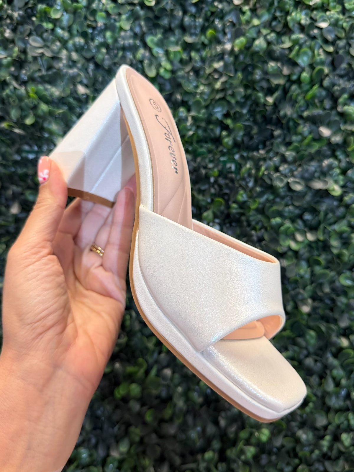White open toe heel held by a hand against a green leafy background