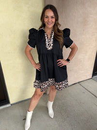 A woman modeling a black short-sleeve dress with leopard print trim on the neckline and hem, paired with white ankle boots, standing against a beige stucco wall.