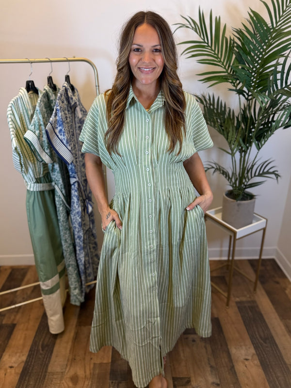 Woman wearing a short sleeve green striped dress with button front and collared neck standing in a room with clothing racks and plants.