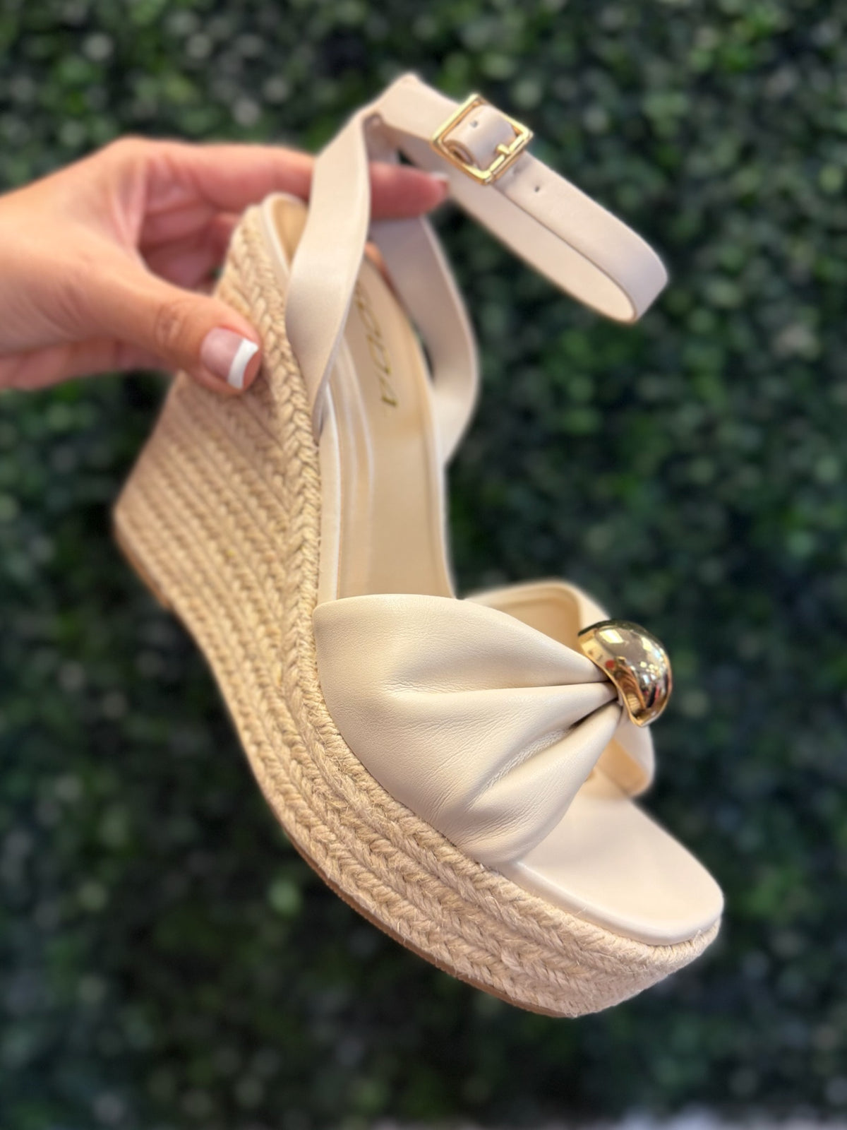 Beige wedge sandal with a bow and gold buckle held against a green leafy background