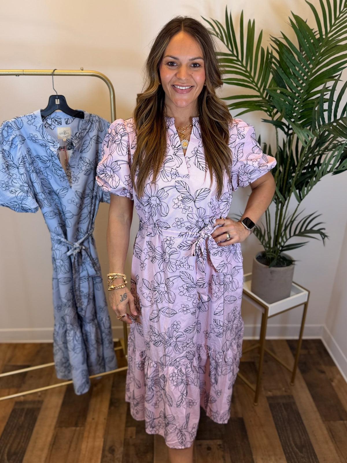 Woman wearing a short sleeve pink midi dress with navy floral embroidery and adjustable waist tie detail standing next to a blue dress on a rack in a room with a plant.