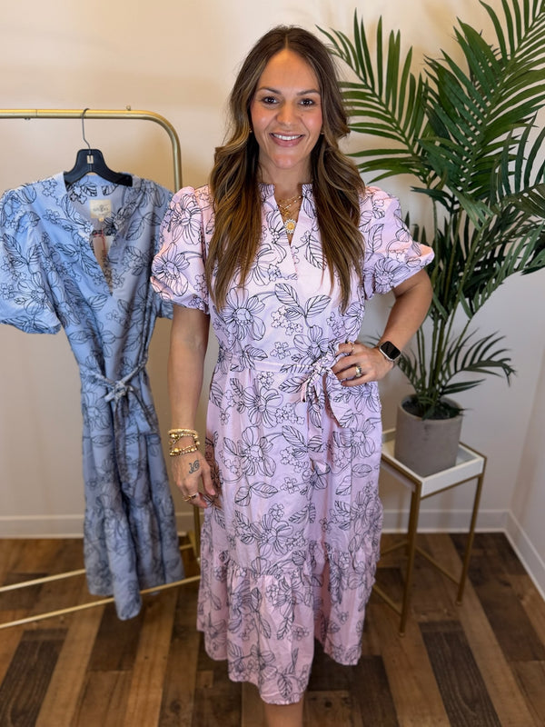 Woman wearing a short sleeve pink midi dress with navy floral embroidery and adjustable waist tie detail standing next to a blue dress on a rack in a room with a plant.
