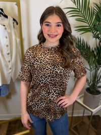Person wearing a leopard print top in a room with plants and clothing.