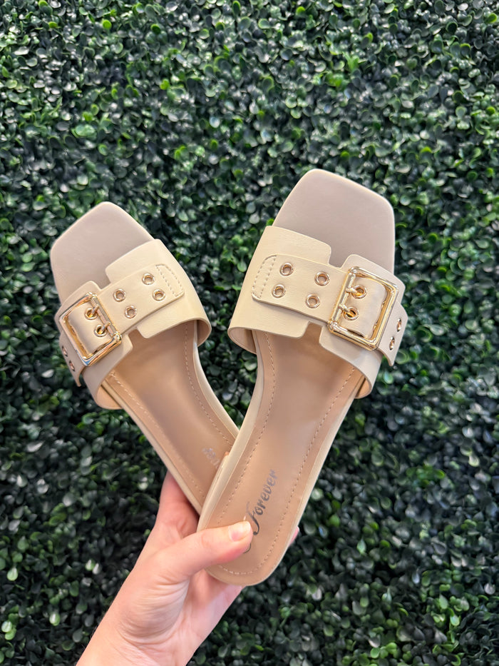 Beige sandals with gold buckles held against a green textured background