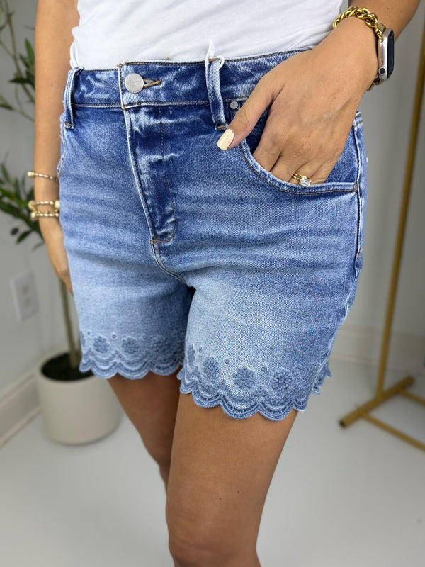 Woman wearing medium wash high rise denim shorts with a scalloped trim and embroidery standing in a fitting room.
