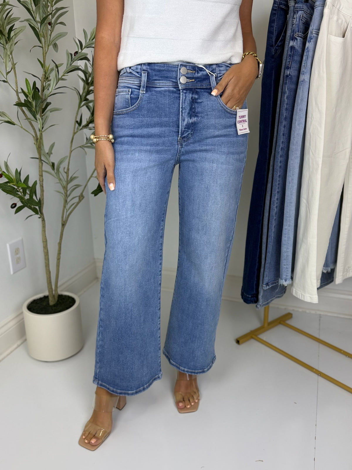 Person wearing medium wash blue jeans with double button waistband detail standing in a room with a clothing rack and plant in the background