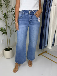 Person wearing medium wash blue jeans with double button waistband detail standing in a room with a clothing rack and plant in the background