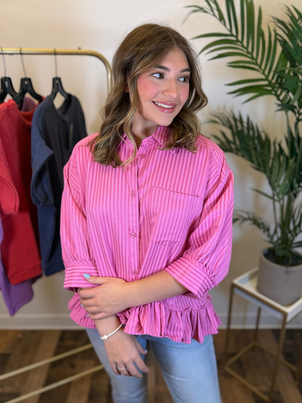 A woman wearing a pink and white vertically striped button-down shirt with a ruffled hem, paired with light blue jeans, standing in a boutique setting.
