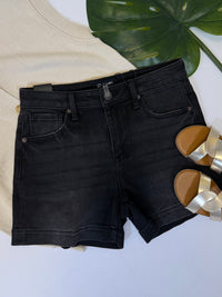 Black denim shorts on a white surface with a beige top and gold sandals in the background.