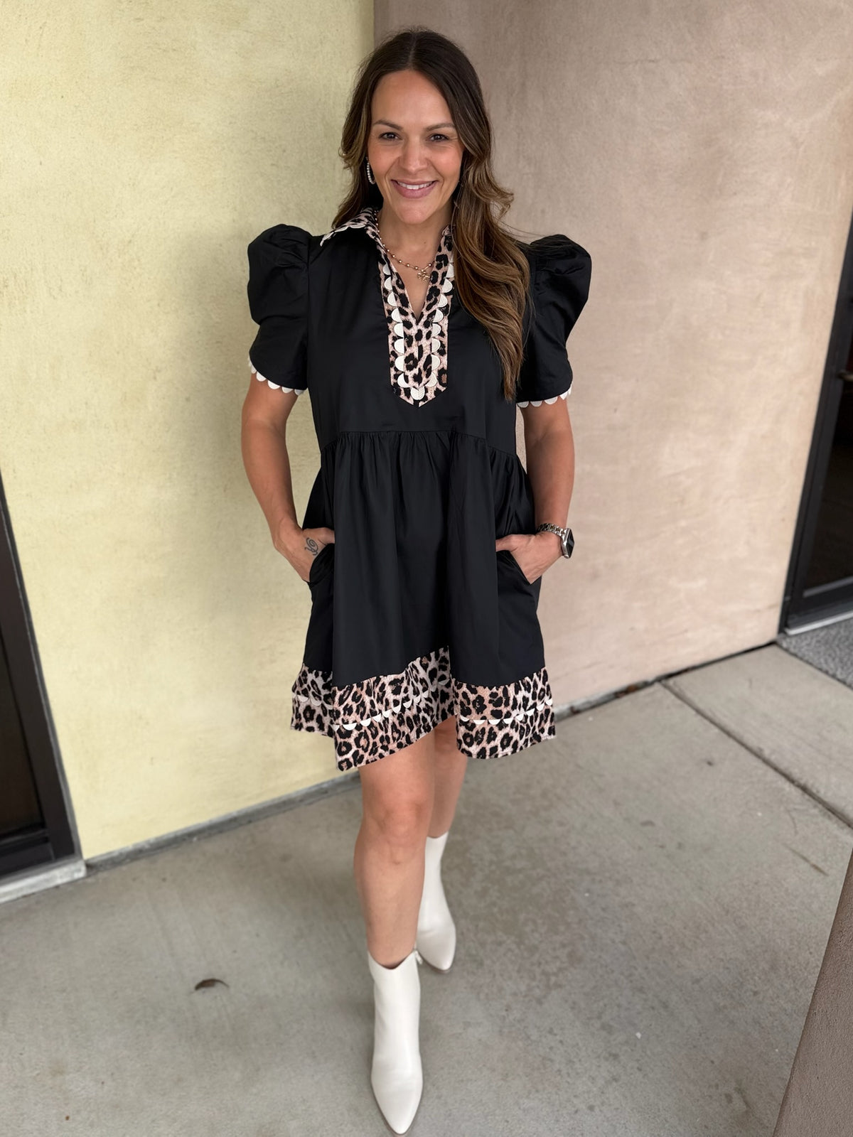 A woman modeling a black puff-sleeve mini dress with leopard print trim at the neckline and hem, paired with white pointed-toe boots, standing against a beige stucco wall.
