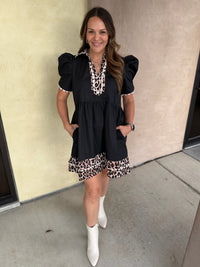 A woman modeling a black puff-sleeve mini dress with leopard print trim at the neckline and hem, paired with white pointed-toe boots, standing against a beige stucco wall.