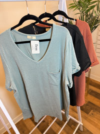 Light sage ribbed v-neck top with rolled sleeves and pocket, hanging with black and rust tops.