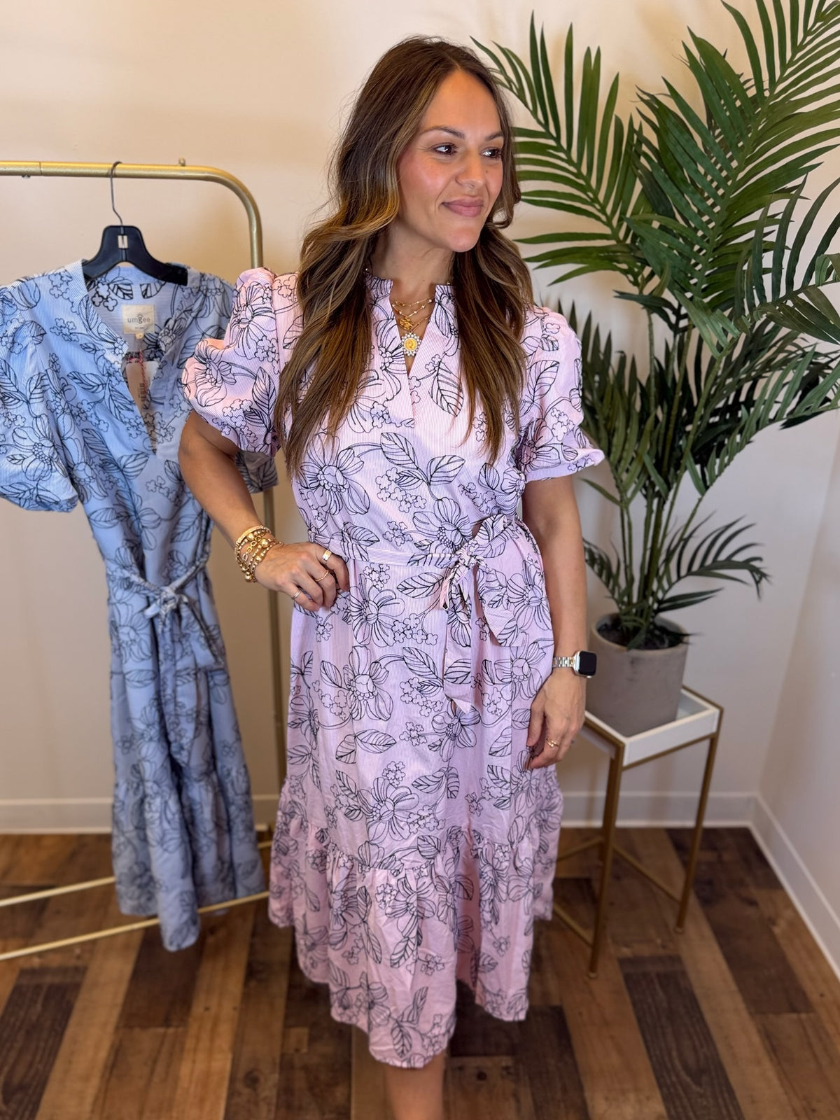 Woman wearing a pink midi dress with navy floral embroidery and split neckline standing next to a rack of dresses in a room with a plant.