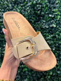 Beige slip on sandal with a gold buckle held against a green leafy background