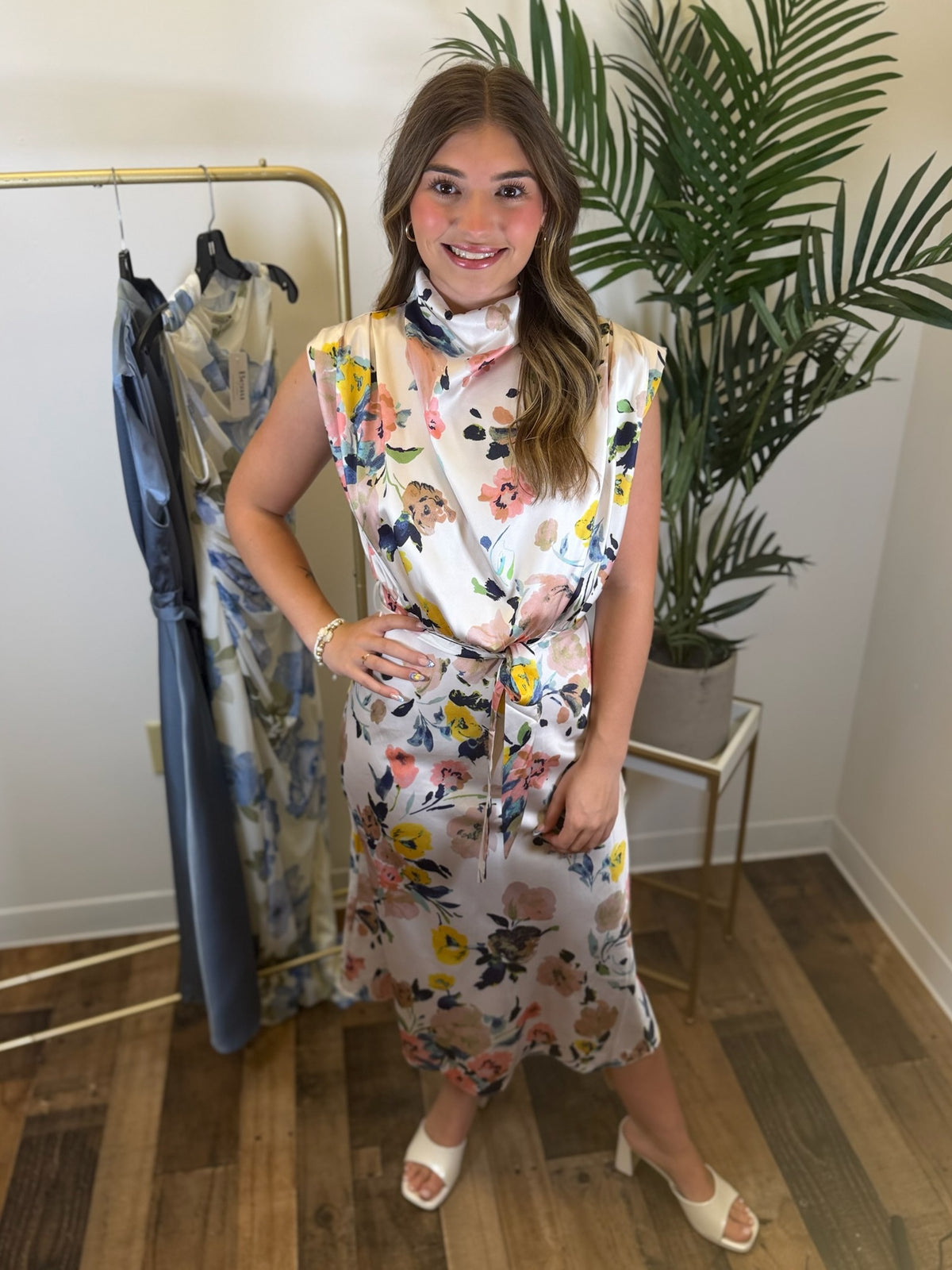 Woman wearing a sleeveless multicolor floral midi dress with draped neckline and adjustable waist details standing in a room with a plant and clothing rack.