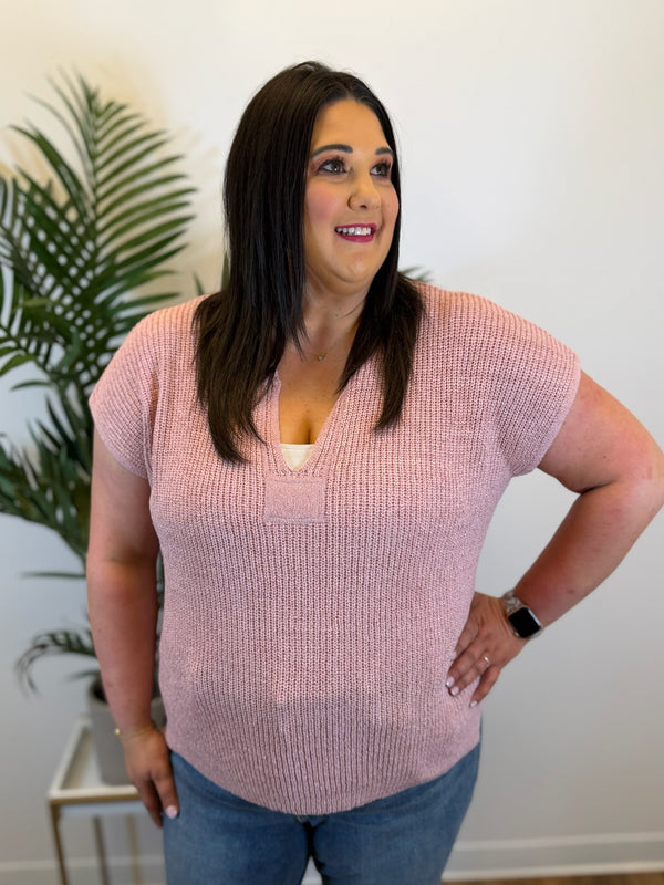 Woman wearing a pink short-sleeve sweater standing indoors with a plant in the background