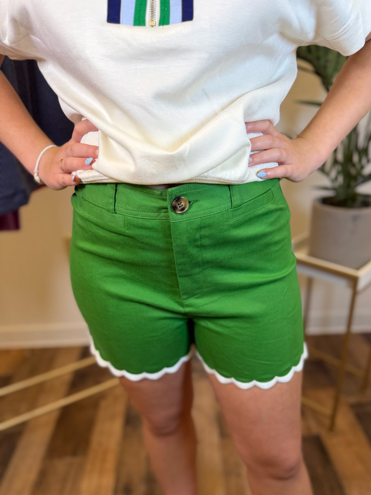 A person wearing bright green shorts with a white scalloped hem and a white top, standing indoors with hands on hips.