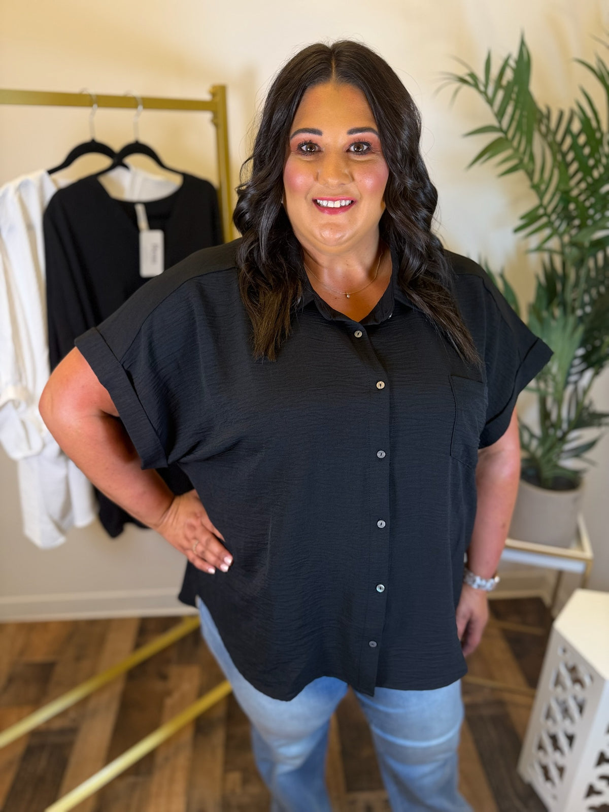 A woman modeling a black short-sleeve button-up shirt with light blue jeans, standing in a boutique setting with a clothing rack and potted plant in the background.